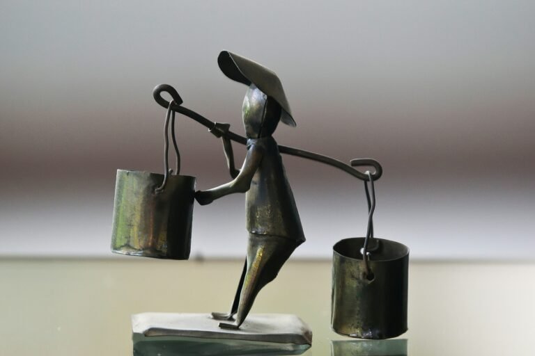 Metal figurine of person carrying to unevenly balanced buckets across their shoulder and struggling