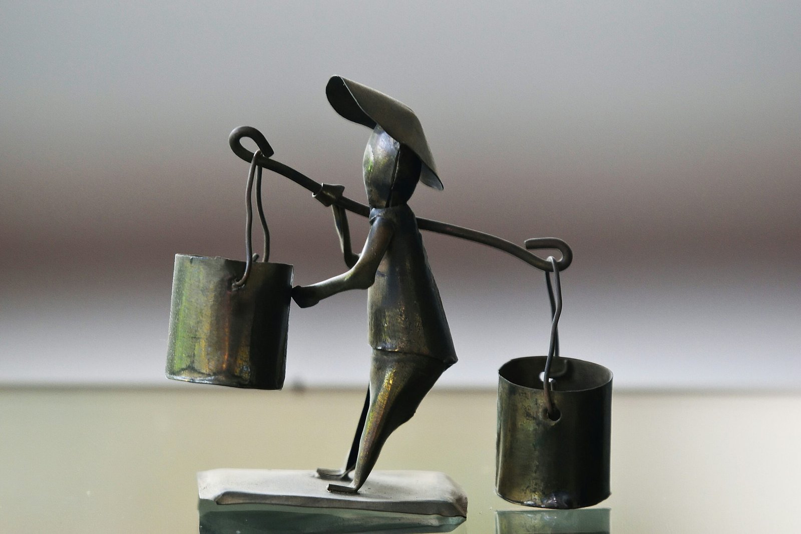 Metal figurine of person carrying to unevenly balanced buckets across their shoulder and struggling