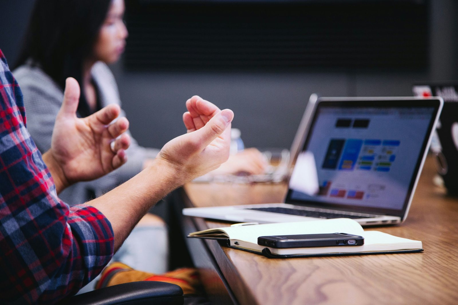 Picture of a man's hands explaining something with a phone, notebook, and computer in background and a blurry woman glancing at the computer